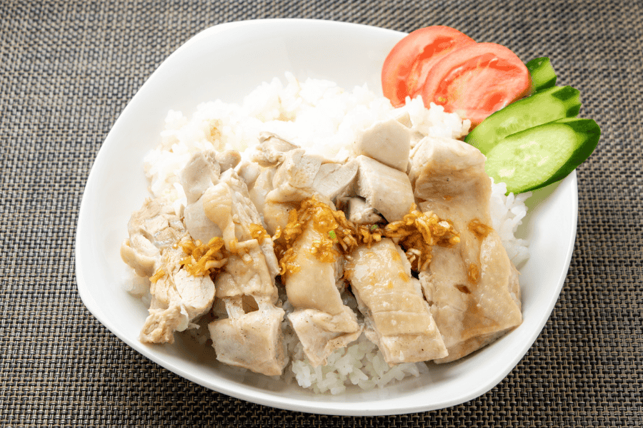 A white bowl of poached chicken slices served over white rice, topped with a savory minced ginger and garlic sauce. The dish is garnished with fresh tomato slices and cucumber rounds on the side.
