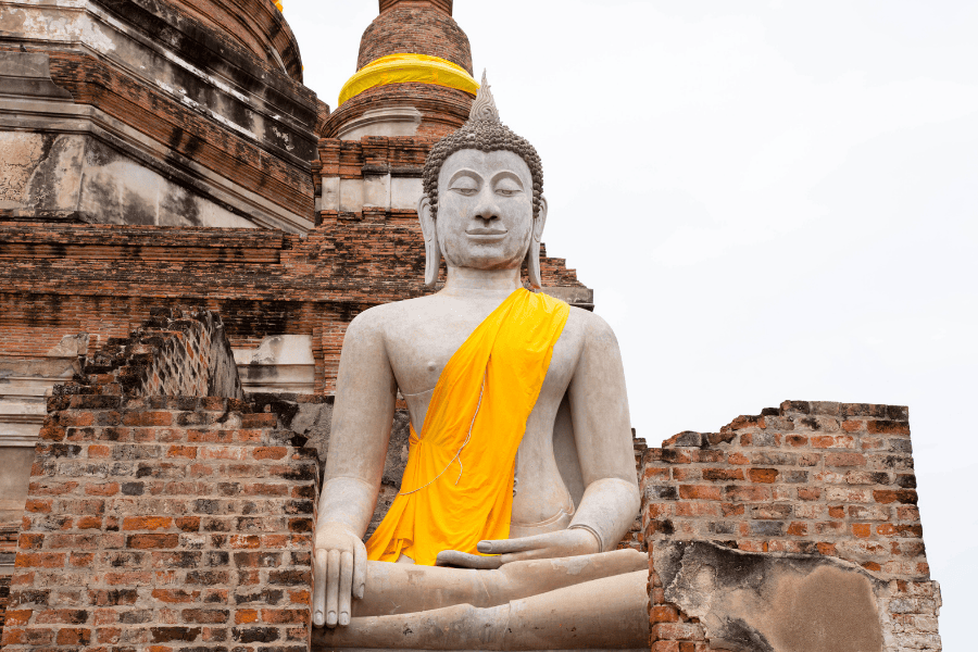 A large, seated stone Buddha statue draped in a bright yellow sash. The statue is positioned in front of the ancient, weathered brick ruins of a stupa at a temple in Ayutthaya, Thailand.