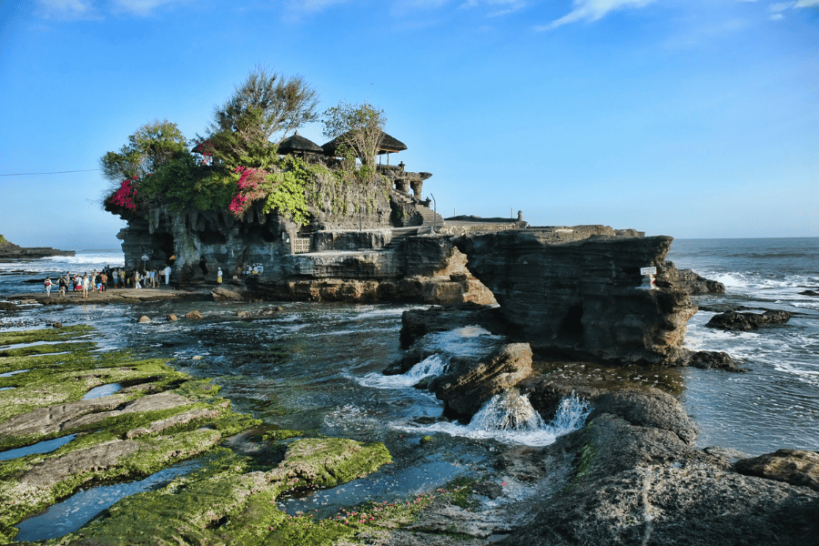 The ancient Hindu pilgrimage temple Tanah Lot sitting atop a large rock formation offshore, with green moss-covered rocks and crashing waves in the foreground under a clear blue sky.