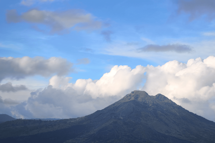 A large, jagged grey volcanic mountain peak rising against a bright blue sky filled with large, fluffy white clouds.