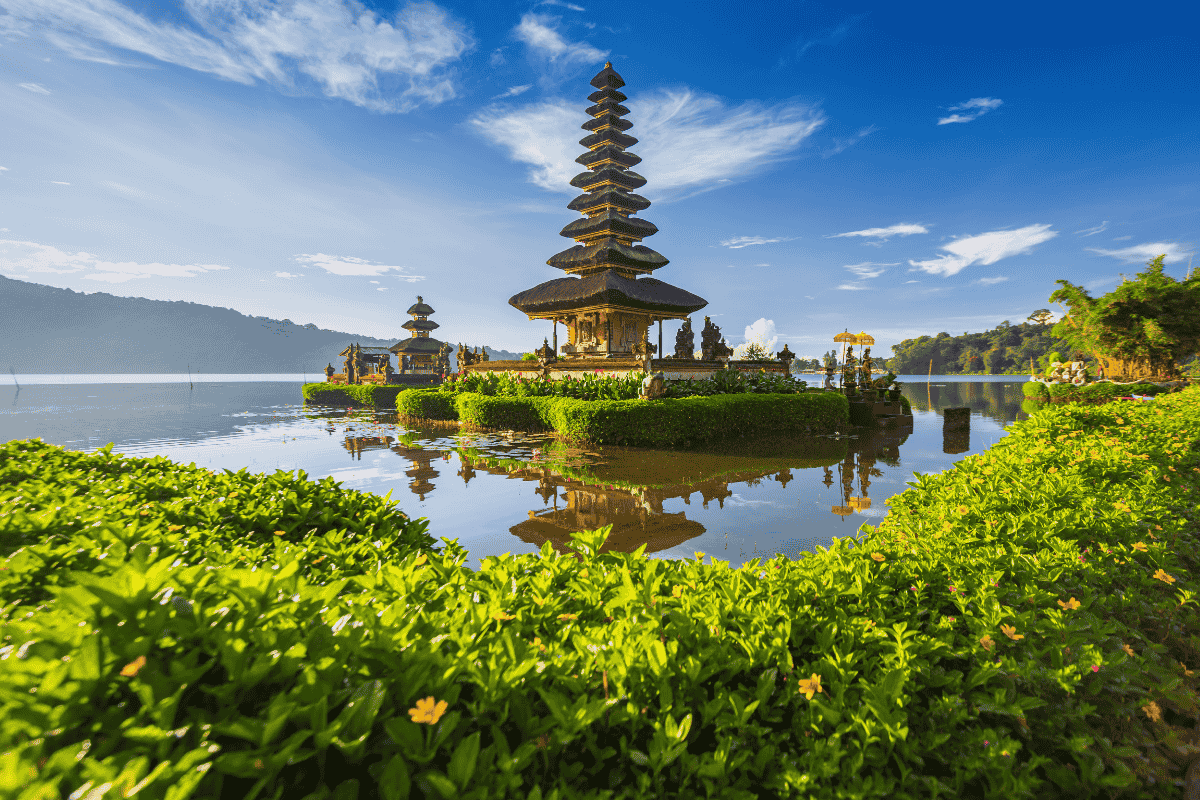 An iconic multi-tiered Hindu water temple, Ulun Danu Beratan, reflecting in a calm lake under a bright blue sky with wispy clouds, framed by lush green gardens in the foreground.