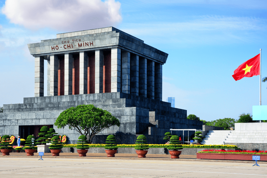 The imposing grey stone Ho Chi Minh Mausoleum in Hanoi, set against a bright blue sky with manicured gardens in the foreground.