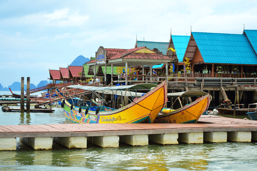 Traditional colorful Thai longtail boats moored at a wooden pier in a floating village with turquoise-roofed stilt houses and limestone karsts in the background.