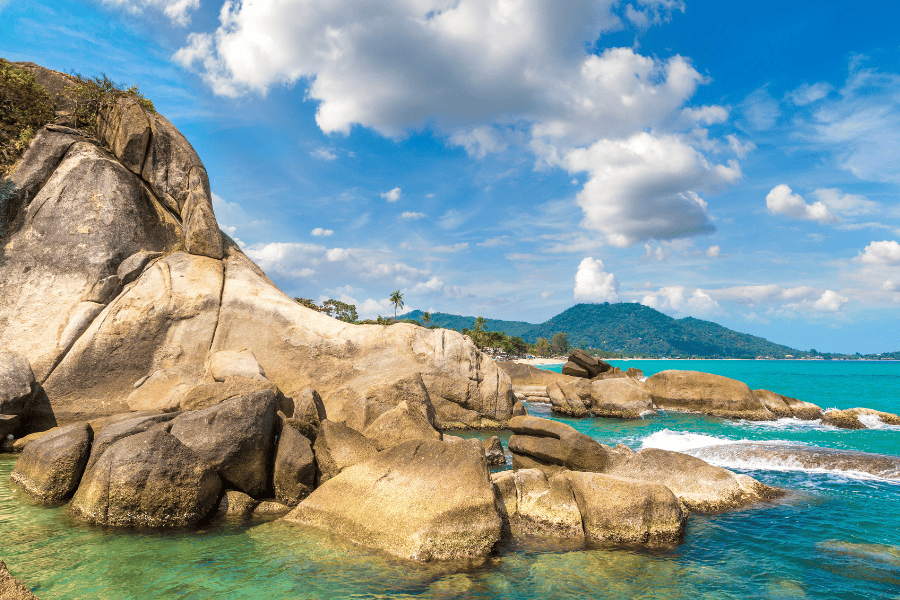 A scenic tropical coastline in Thailand featuring large, smooth tan boulders in the foreground and turquoise ocean water