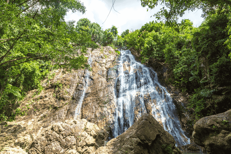 A tall tropical waterfall cascading over a rocky cliff face surrounded by lush green jungle foliage under a bright sky.