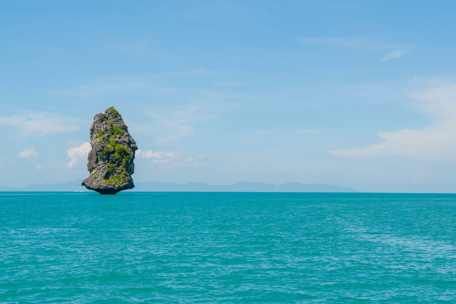 A large, solitary limestone karst island with green vegetation rising from vibrant turquoise waters under a clear blue sky in Thailand.
