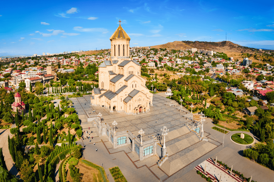 An aerial panoramic view of the massive Holy Trinity Cathedral of Tbilisi (Sameba) with its golden dome, set against a backdrop of the city and rolling hills under a clear blue sky.