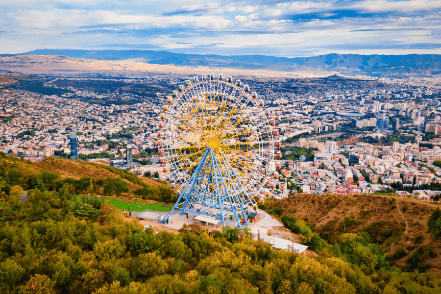 A high-angle panoramic view of a large white and yellow Ferris wheel situated on a lush green hilltop overlooking the sprawling city of Tbilisi, Georgia, with distant mountains under a blue sky.