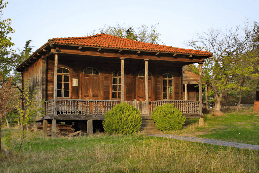 Traditional rustic wooden cottage with a red clay tile roof and a wrap-around veranda in a rural landscape.