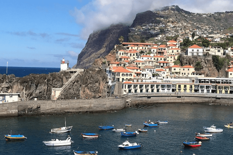 A scenic coastal village with terracotta-roofed white houses built into a steep hillside overlooking a bright blue harbor. Several small fishing boats in blue, white, and red are anchored in the calm water.