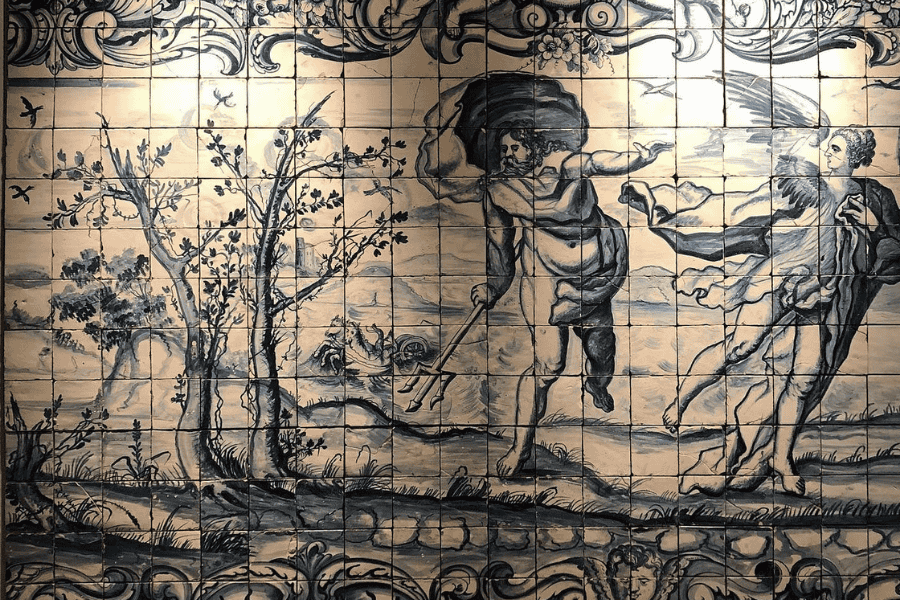A large, detailed azulejo tile mural inside the National Tile Museum in Lisbon, featuring classical blue-and-white hand-painted ceramics. The artwork depicts a mythological scene set in a coastal landscape with thin trees and a distant castle. At the center, a muscular figure—likely Neptune—strides forward carrying a trident, while a winged figure in flowing robes appears to the right. The mural is framed by ornate scrollwork and floral borders, characteristic of 18th-century Portuguese tilework.