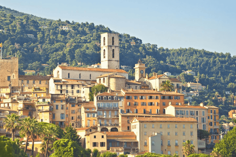 A warm, sunlit view of a hillside town in Grasse, France, featuring a cluster of traditional ochre and cream-colored buildings. The town is dominated by the tall, square stone bell tower of the Grasse Cathedral and a nearby smaller clock tower. Lush green trees and forested hills rise steeply behind the architecture, and palm trees are visible in the foreground, capturing the classic atmosphere of the French Riviera hinterland.