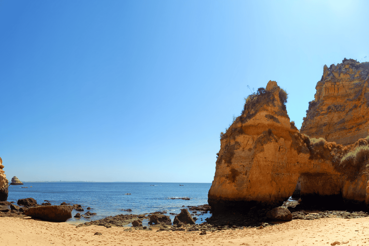 A wide-angle landscape of a golden sandy beach in Lagos, Portugal, featuring dramatic limestone sea stacks and natural rock arches.