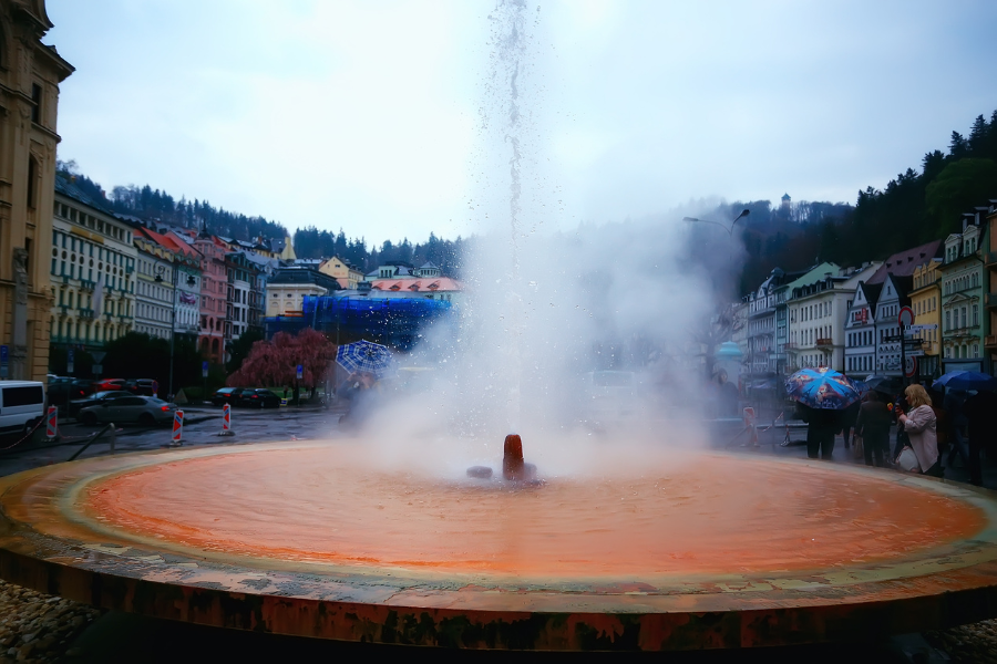 A powerful steaming geyser (the Vřídlo) erupting from a large, circular orange-tinted basin in a town square in Karlovy Vary.
