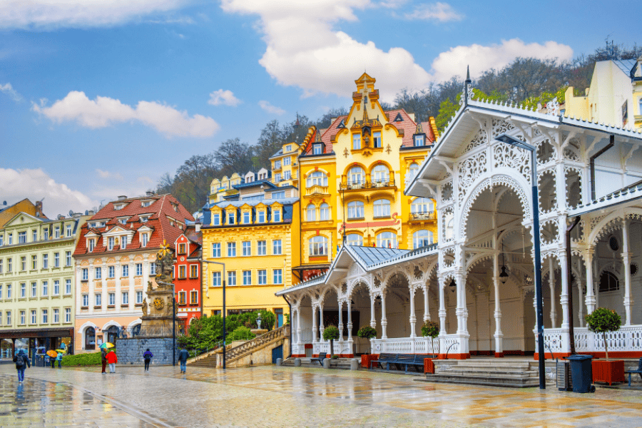 A vibrant town square in Karlovy Vary, Czech Republic, featuring the ornate, white-carved Market Colonnade in the foreground. Behind it stands a row of grand, multi-story buildings in bright yellow and orange with classic European gables.