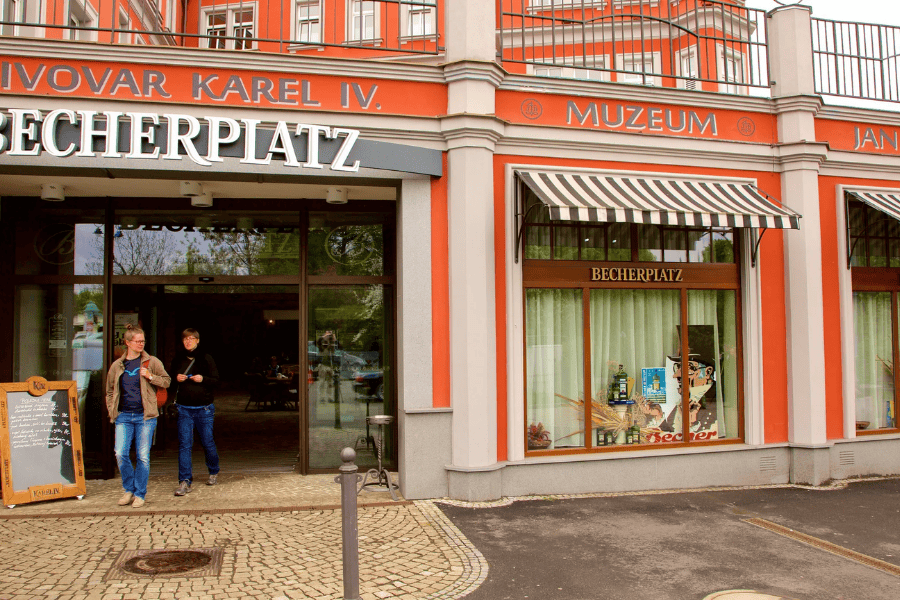 An eye-level shot of the Becherplatz entrance in Karlovy Vary, featuring a vibrant orange facade with grey architectural trim. Two people walk out of the dark glass entrance next to a wooden menu board.