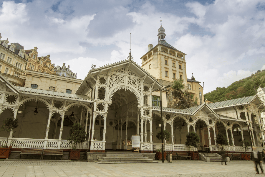 An eye-level, medium shot shows the Market Colonnade in Karlovy Vary, Czech Republic. The white, wooden structure features intricate fretwork and Swiss-style architecture.