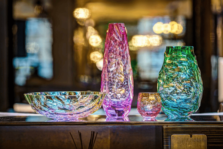 A collection of Bohemian glassware displayed on a reflective surface. The set includes a shallow iridescent bowl, a tall pink textured vase, a small round pink glass, and a green textured vase.